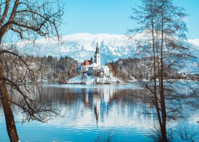 An island on Lake Bled covered in snow, snowy Alps in the background.