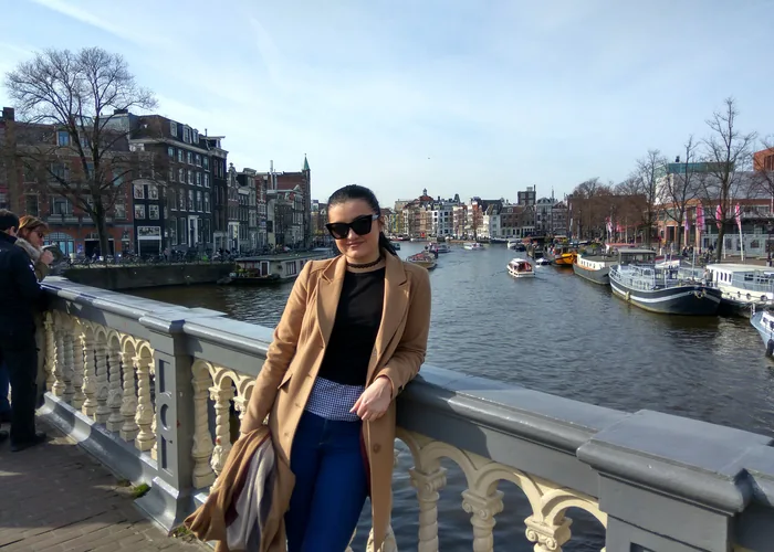 Ana stoji na mostu s brodicama i kućama u pozadini u Amsterdamu.