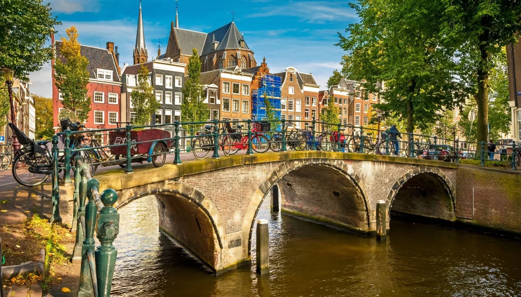 Bicikli parkirani na mostu iznad kanala, povijesne kuće i crkva u Amsterdamu.